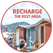 The image displays three wooden small houses alternating between maroon and light blue. The roofs are made of metal and wood. The houses are being built in the photo, and groups of people are scattered around with long pieces of wood and toolboxes. There is red text at the top of the page that reads "RECHARGE THE REST AREA." 