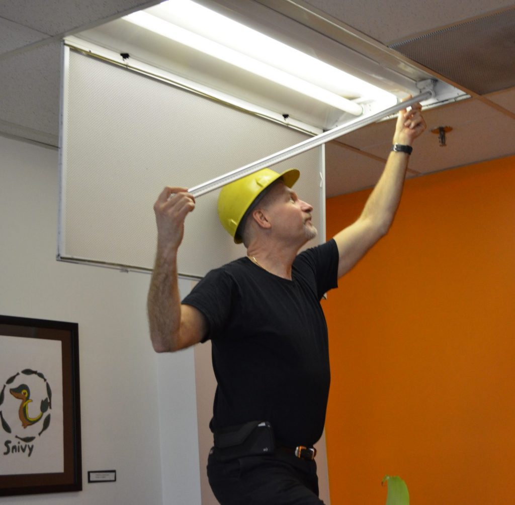 A man wearing a safety helmet install a LED light overhead in a room with an orange wall.