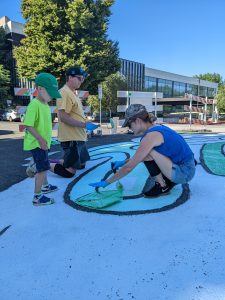 Two adults and one child are painting a street with green paint over a white primer paint base coat.