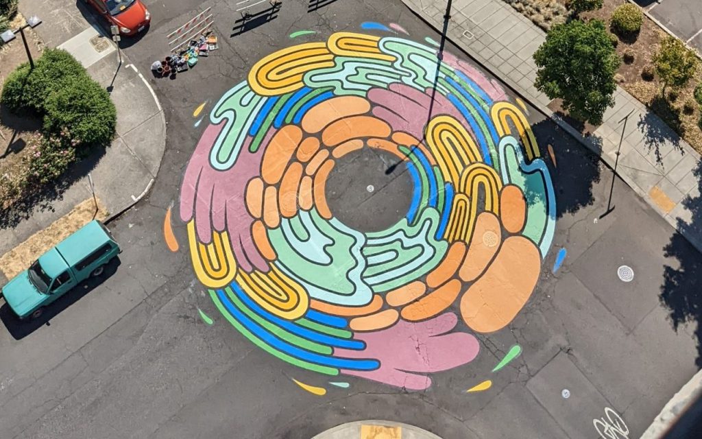 The image is of a mural on the ground that is a symbol of the intersection repair project. The mural is an abstract circle that shows different colors with lines running through them. Some of the colors are forest green, olive green, pastel purple, bright yellow, and red.