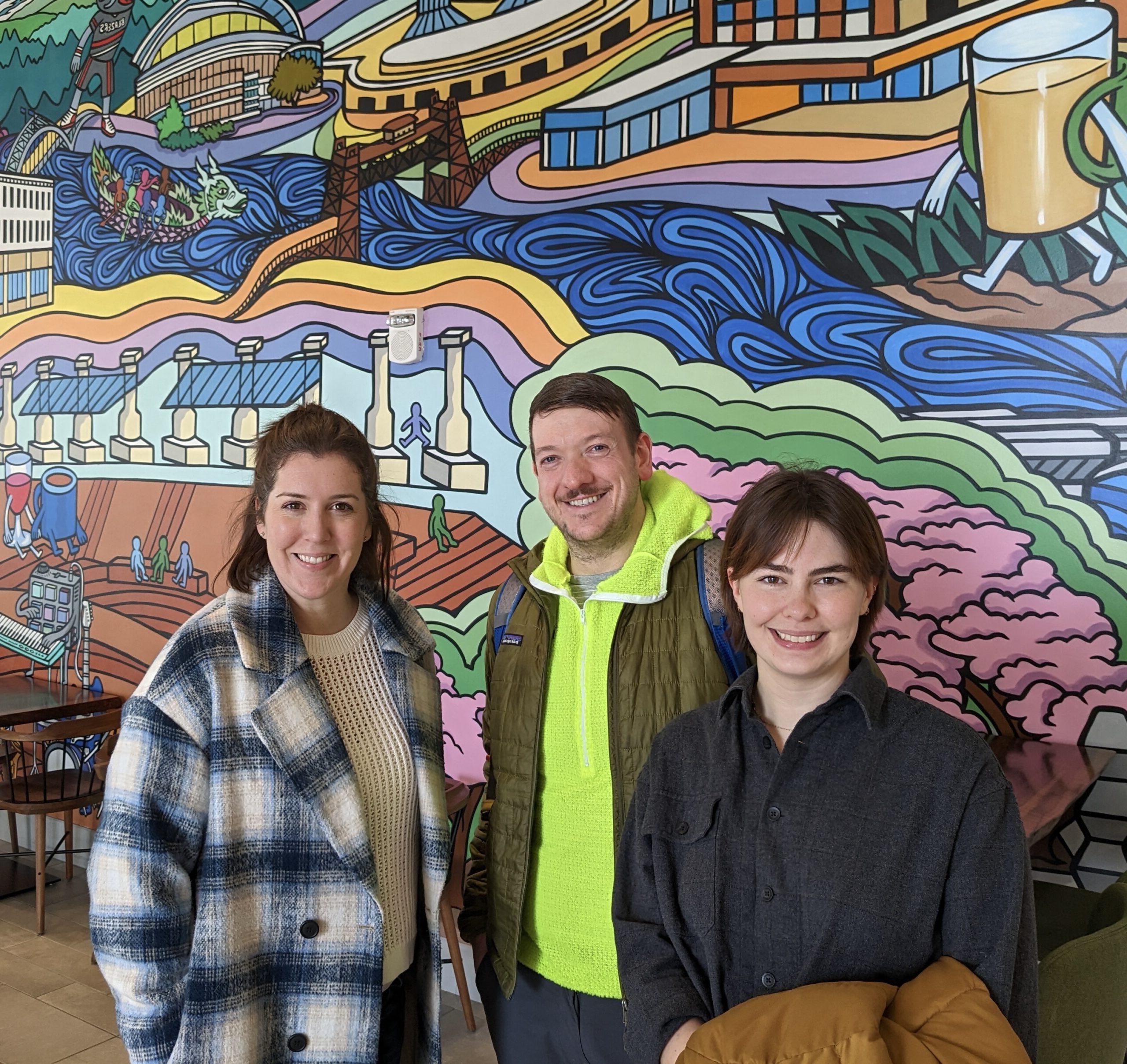 Three Lloyd EcoDistrict staff members stand in front of a colorful mural. From left to right: Kristin Leiber, Joshua Baker, and Burgin Utaski.