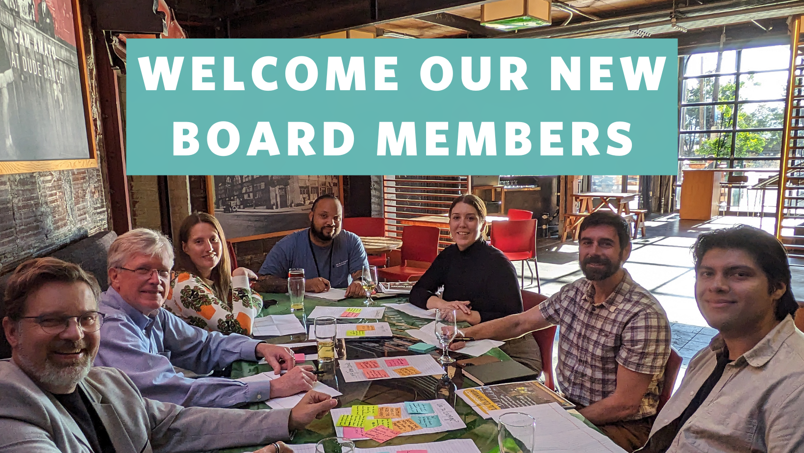 Photo of 7 people sitting at a table looking at the camera smiling with the words “welcome our our new board members” overlaying the image