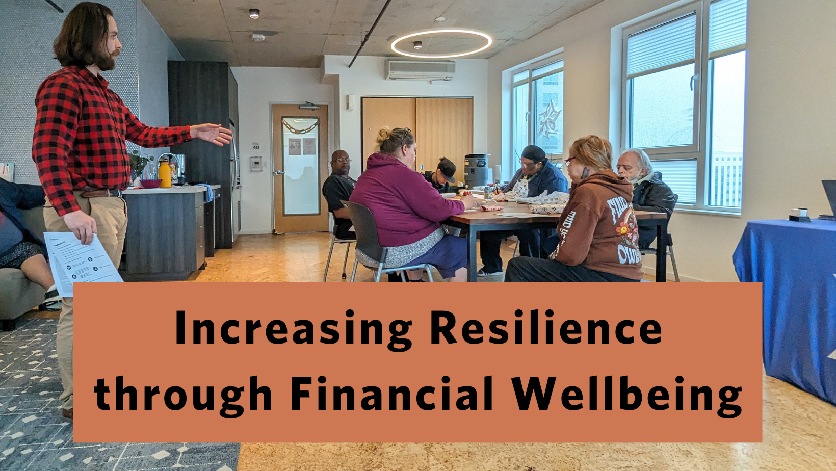 a picture of 6 people sitting around a table listening to a man standing up presenting with the words “Increasing Resilience through Financial Wellbeing”.