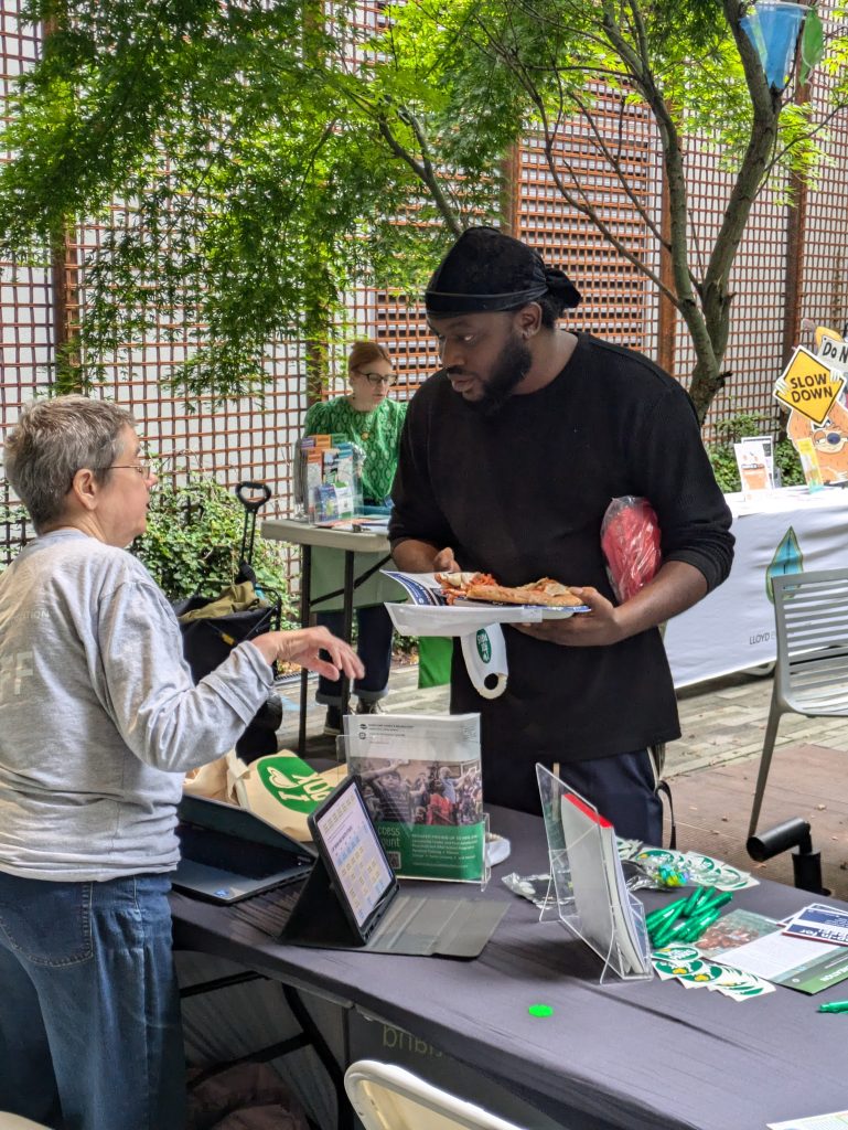 An outdoor photo of a tabling event. Someone holding a plate of food is talking to the person behind the table, who is assisting them with something.