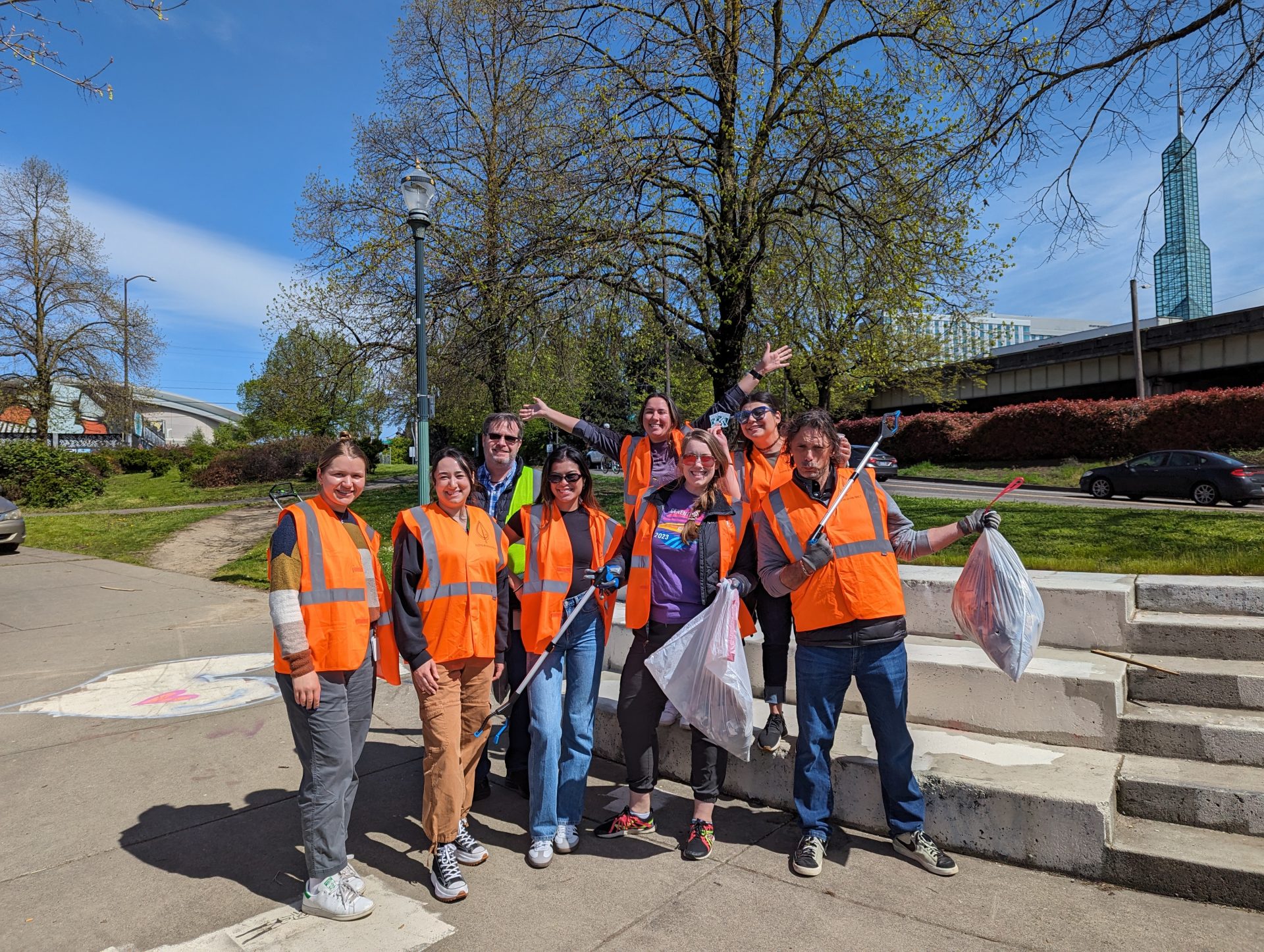 image of a eight volunteers posing a a camera at Peace Memorial park. They are wearing orange safety vests and hold litter grabbers or garbage bags.