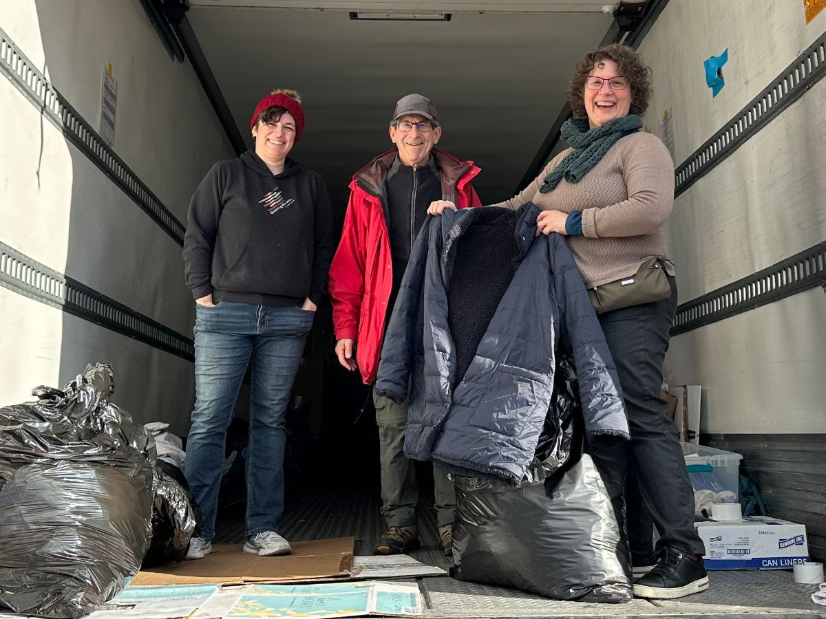 3 people in the back of a semi truck, with the person on the right holding up a winter coat.