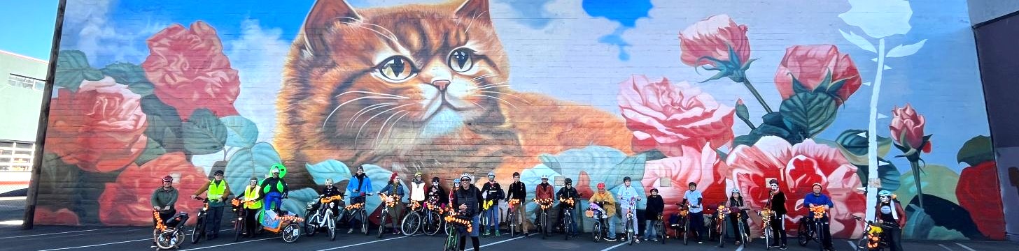 a group of riders with marigolds on their bikes pose Infront of a kitten mural