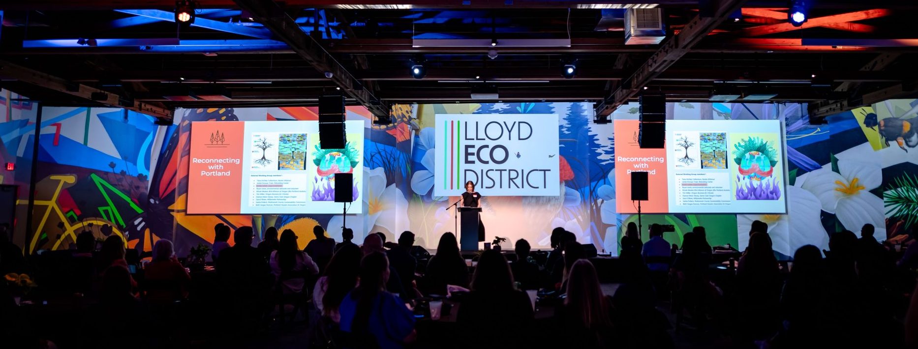image of kristin leiber on stage at model the way 2024 with artwork and LLoyd EcoDistrcit logo projected on the walls