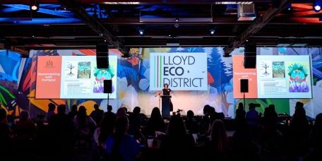 image of kristin leiber on stage at model the way 2024 with artwork and LLoyd EcoDistrcit logo projected on the walls