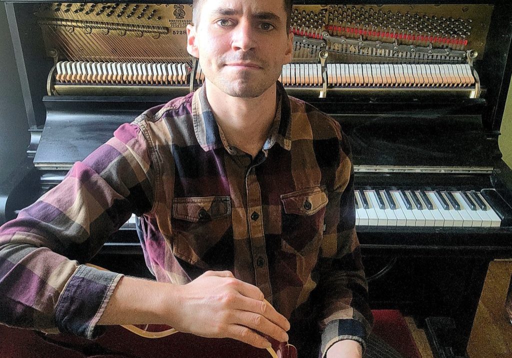 Ben Woodman with a guitar, piano in the background