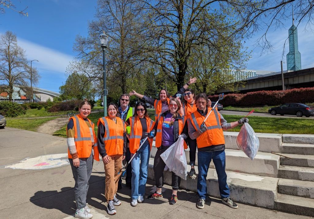 image of a eight volunteers posing a a camera at Peace Memorial park. They are wearing orange safety vests and hold litter grabbers or garbage bags.