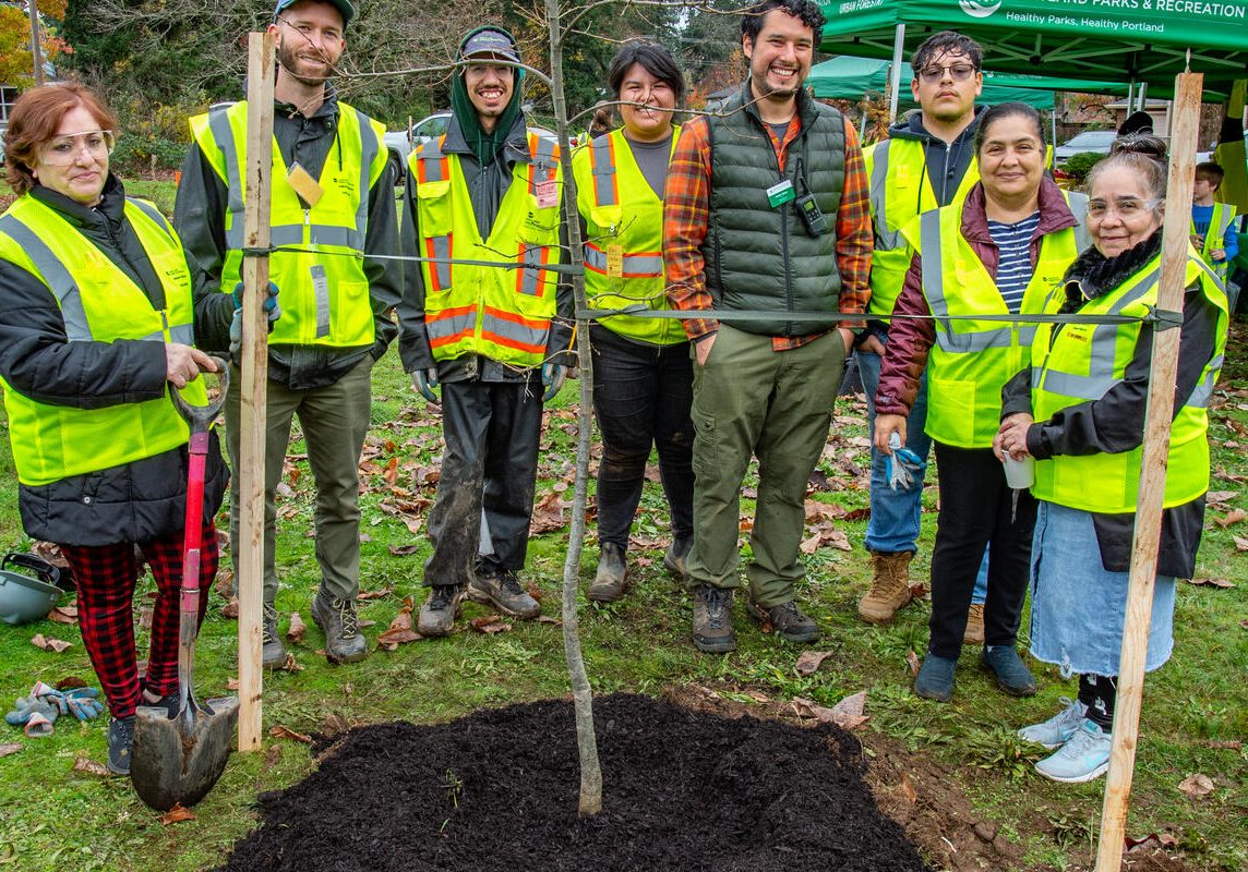 a group of people in neon green safety vests pose of a photo behind a newly planted tree.