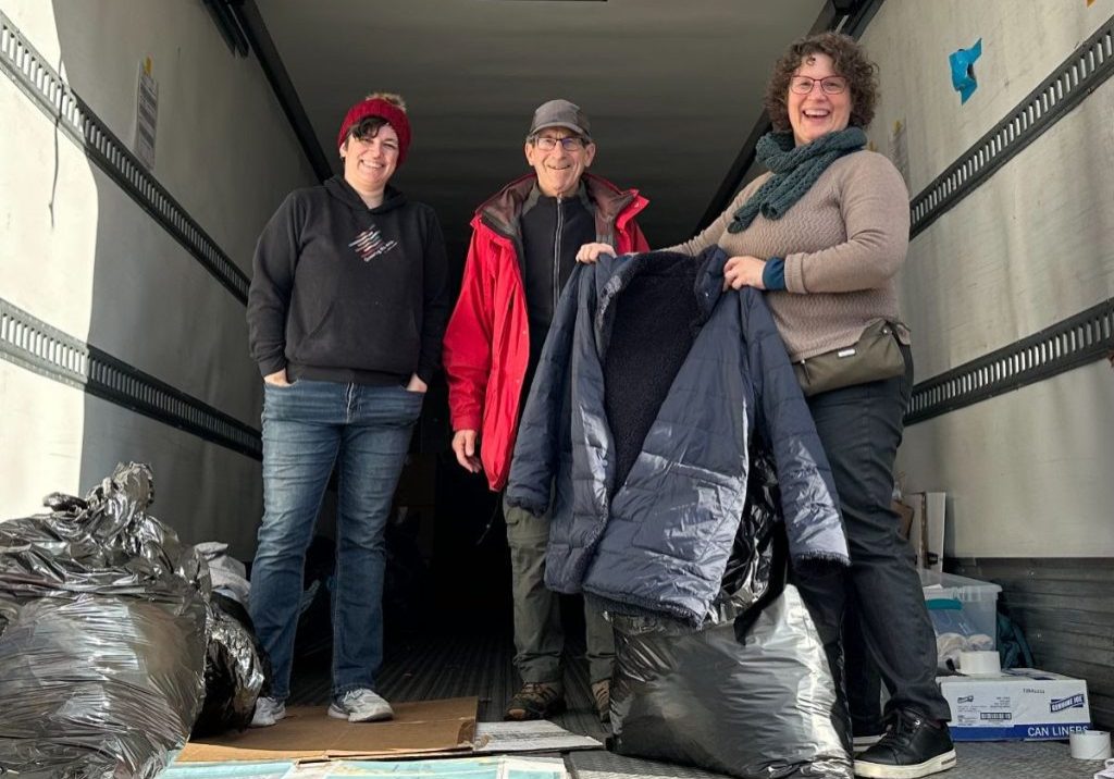 3 people in the back of a semi truck, with the person on the right holding up a winter coat.