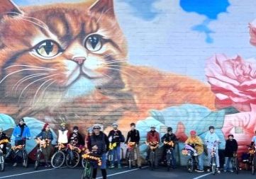 a group of riders with marigolds on their bikes pose Infront of a kitten mural